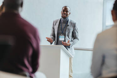 Professional male speaker with a beaming smile engages an audience at a business seminar. The conference room is filled with natural light, emphasizing a positive and productive atmosphere. - JLPSF31406