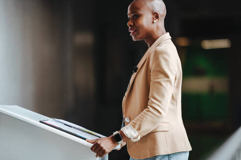 Eine elegante Frau steht selbstbewusst da und hält eine Geschäftsrede. Mit ihrem eleganten Blazer und ihrer schicken Kleidung strahlt sie Professionalität aus und ist damit die ideale Verkörperung von Führung und Erfolg am Arbeitsplatz. - JLPSF31414