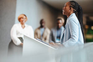 Eine erfolgreiche afrikanische Geschäftsfrau unterhält sich mit ihren Zuhörern, während sie auf einer Fachkonferenz eine inspirierende Rede hält. Die Kollegen hören in einer modernen Unternehmensumgebung aufmerksam zu. - JLPSF31427