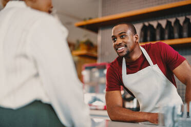 Ein fröhlicher männlicher Barista in einer Schürze bietet exzellenten Kundenservice in einem modernen Café. Das helle Interieur und die angenehme Interaktion stellen eine warme, einladende Atmosphäre dar, die perfekt für Lifestyle- und Gastgeberthemen ist. - JLPSF31436