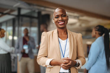 Eine strahlende, professionelle afroamerikanische Frau steht selbstbewusst lächelnd in einer Geschäftsumgebung, während ihre Kollegen im Hintergrund ein Gespräch führen. Sie strahlt Führung und Positivität aus. - JLPSF31449