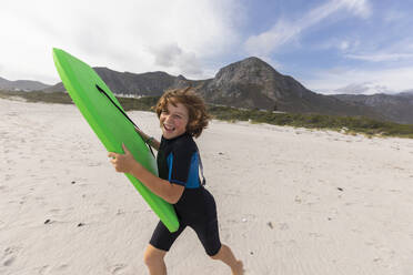 Südafrika, Hermanus, Lächelnder Junge läuft am Strand mit Bodyboard - TETF02578