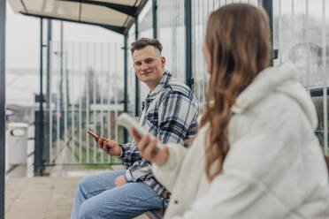 Young couple sitting on bench holding smart phones at train station - VSNF01702