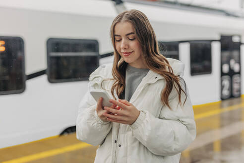 Smiling woman using smart phone at railroad station - VSNF01703