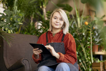 Smiling gardener sitting with tablet PC at store - SANF00194