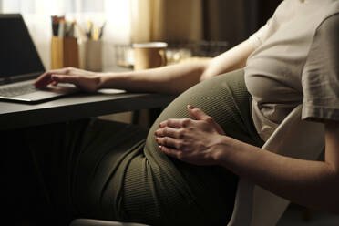 Schwangere Frau berührt ihren Bauch, während sie auf einem Stuhl sitzt - DSHF01617