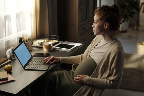 Schwangere junge Frau arbeitet am Laptop am Schreibtisch im Heimbüro - DSHF01625