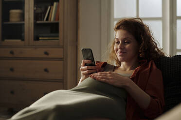 Smiling pregnant woman using smart phone in living room - DSHF01638