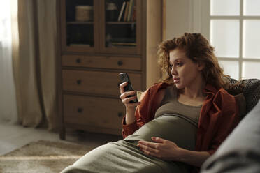 Pregnant woman using mobile phone lying on sofa at home - DSHF01639