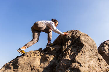 Low angle of cheerful brunet guy in casual outfits climbing rough mountain slope against clear blue sky during sightseeing in Los Dinamos close to Mexico - ADSF53211