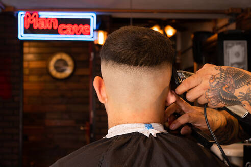 Anonymer tätowierter Friseur, der die Haare eines jungen männlichen Kunden mit einem Trimmer schneidet, während er sich in einem Friseursalon pflegt - ADSF53212