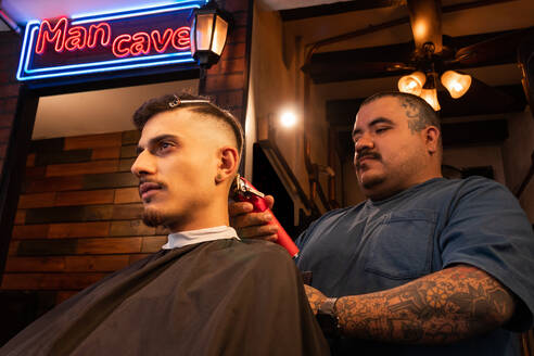 Side view of man in barber cape sitting and getting haircut during grooming routine at barbershop against neon sign and lights on background - ADSF53214