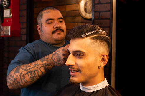 A tattooed barber cutting his hair of a man in a barber's cape sitting and during his grooming routine in a barber's shop - ADSF53215