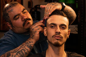 Seriöser Friseur mit tätowiertem Arm schneidet Haare von stilvollem Kerl mit Trimmer während professioneller Pflegeprozess im Friseursalon - ADSF53216