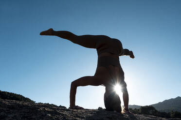 Back view of unrecognizable sportswoman in activewear performing headstand on rough hill slope against blue cloudless sky during fitness training - ADSF53267