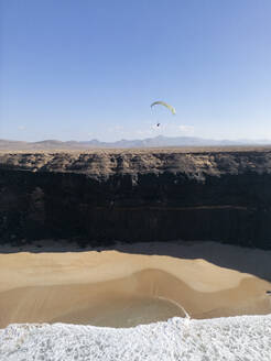 Ein einsamer Gleitschirm, der über die Klippen und Sanddünen von Fuerteventura fliegt, mit dem schäumenden Meer am Ufer und den Bergen in der Ferne - ADSF53271