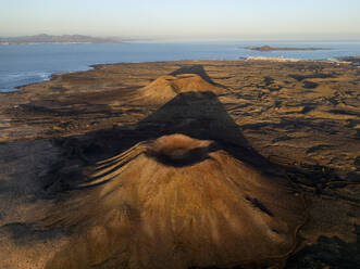 Eine Luftaufnahme zeigt die dramatische vulkanische Landschaft in Küstennähe mit einer kleinen Stadt in der Ferne unter einem goldenen Sonnenuntergangshimmel - ADSF53272