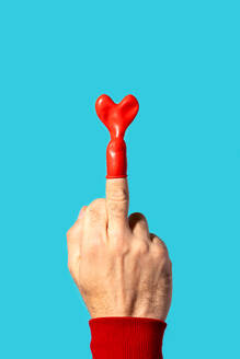 Anonymous person hand in full sleeve sweater and wearing heart shaped red balloon neck on middle finger in bright light against blue background - ADSF53323