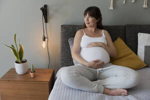 Glückliche schwangere Frau sitzt auf dem Bett und berührt ihren Bauch zu Hause - INGF12904