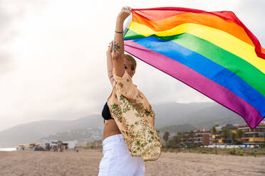 Beautiful lesbian woman holding LGBT flag at the beach, concepts about LGBTQ community, diversity, love and lifestyle - DMDF10348