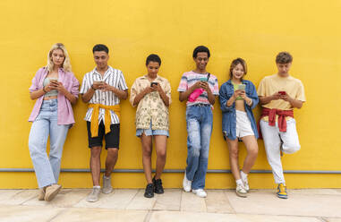Eine Gruppe von Freunden lehnt an einer gelben Wand und benutzt ihre Smartphones - OIPF03923
