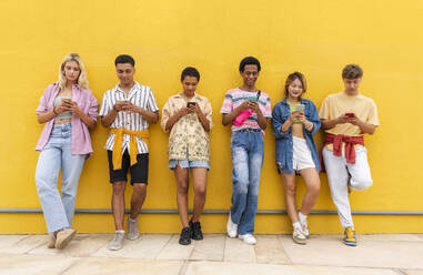 Group of friends leaning against yellow wall using their smartphones - OIPF03923