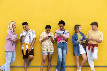 Group of friends leaning against yellow wall using their smart phones - OIPF03927