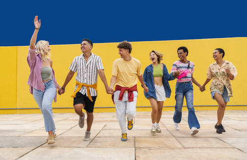 Multi-ethnische Gruppe junger Freunde läuft vor einer gelben Wand und hält sich an den Händen - OIPF03938