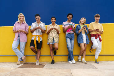 Group of friends leaning against yellow wall using their smartphones - OIPF03952