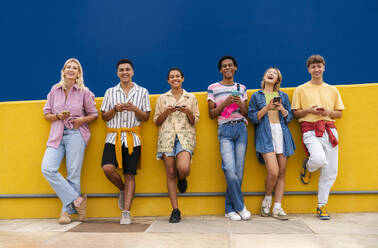 Group of friends leaning against yellow wall using their smartphones - OIPF03953