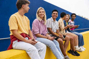 Happy friends with colorful clothing sitting on yellow wall holding smartphones - OIPF03964