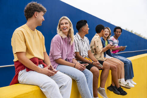 Glückliche Freunde mit bunter Kleidung sitzen an einer gelben Wand und halten Smartphones - OIPF03965