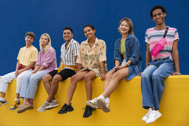 Smiling friends with colorful clothing sitting on yellow wall together - OIPF03966