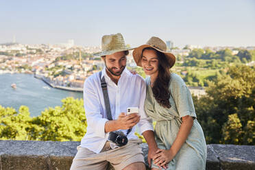 Smiling couple wearing hat and sharing smart phone at Porto city in Portugal - BSZF02630