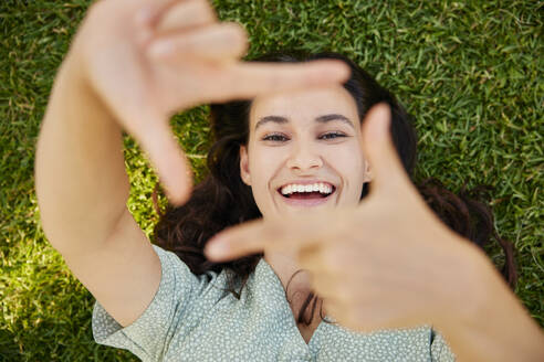 Happy woman looking through finger frame and lying on grass - BSZF02647