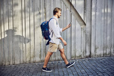 Young man walking with backpack and using smart phone on footpath - BSZF02658