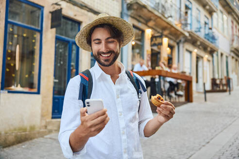 Happy young man holding traditional Portuguese dessert and using smart phone - BSZF02664