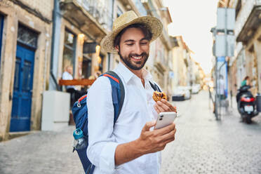 Lächelnder junger Mann mit Smartphone und traditionellem Dessert in der Hand - BSZF02665