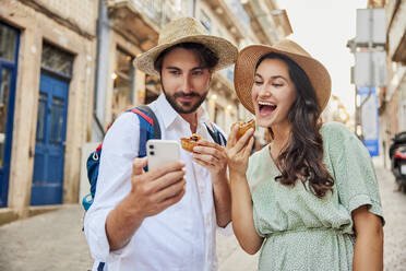 Happy couple taking selfie with traditional dessert pastel de nata through smart phone - BSZF02667