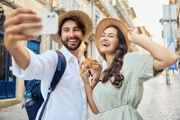 Smiling young couple wearing hat and taking selfie with traditional dessert pastel de nata through smart phone - BSZF02669