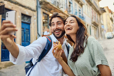 Cheerful couple holding traditional dessert pastel de nata and taking selfie through smart phone - BSZF02670