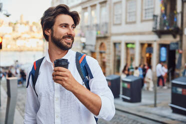 Smiling young man holding coffee cup and standing on street - BSZF02671