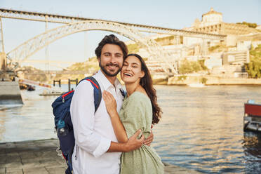 Smiling couple standing near Douro river in front of Dom Luis bridge at Porto, Portugal - BSZF02681