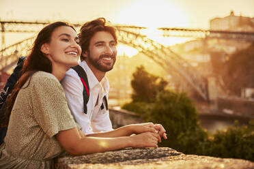 Smiling couple holding hands and standing in front of Dom Luis bridge at sunset, Porto, Portugal - BSZF02705