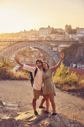 Fröhliches Paar mit erhobenen Armen vor der Dom-Luis-Brücke bei Sonnenuntergang, Porto, Portugal - BSZF02707