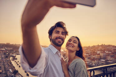 Happy couple taking selfie through smart phone in front of Porto city at sunset, Portugal - BSZF02719