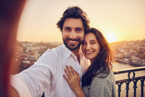 Happy loving couple taking selfie through smart phone in front of Porto city at sunset, Portugal - BSZF02720