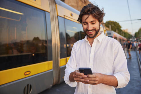Fröhlicher junger Mann benutzt sein Smartphone in der Nähe der Straßenbahn - BSZF02721