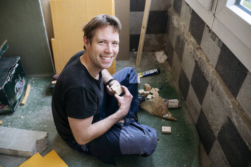 Lächelnder Mann, der auf dem Boden sitzt und sein Haus renoviert - NJAF00806