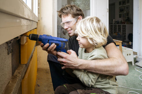 Smiling father teaching boy to drill wall at home - NJAF00819